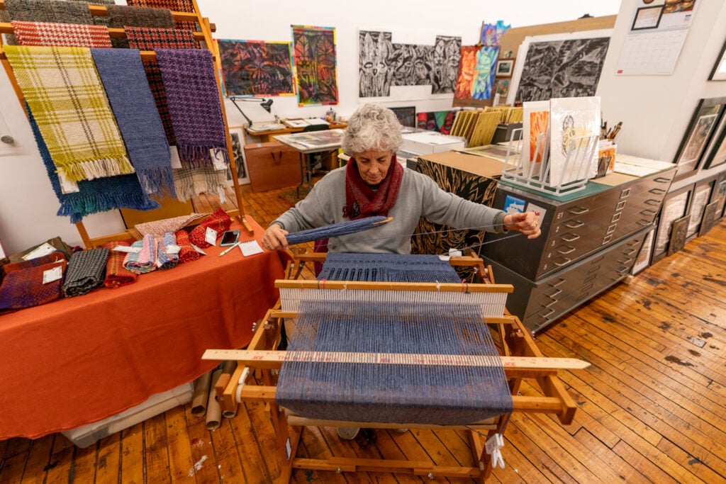 A woman is weaving a blue yarn through a loom inside of a shared studio space at the Hatch Street Open Studios in the spring.